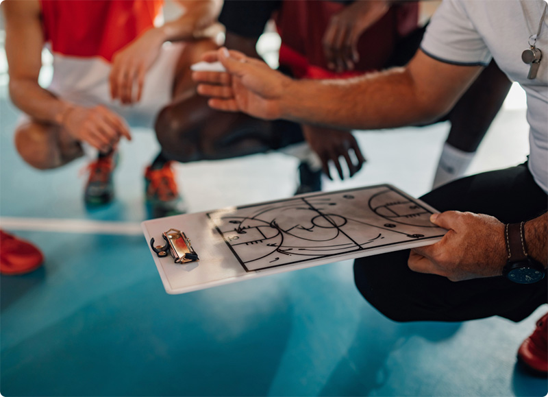 A basketball coach holds a clipboard with a court diagram, explaining strategy to players who are seated and listening intently on an indoor court.