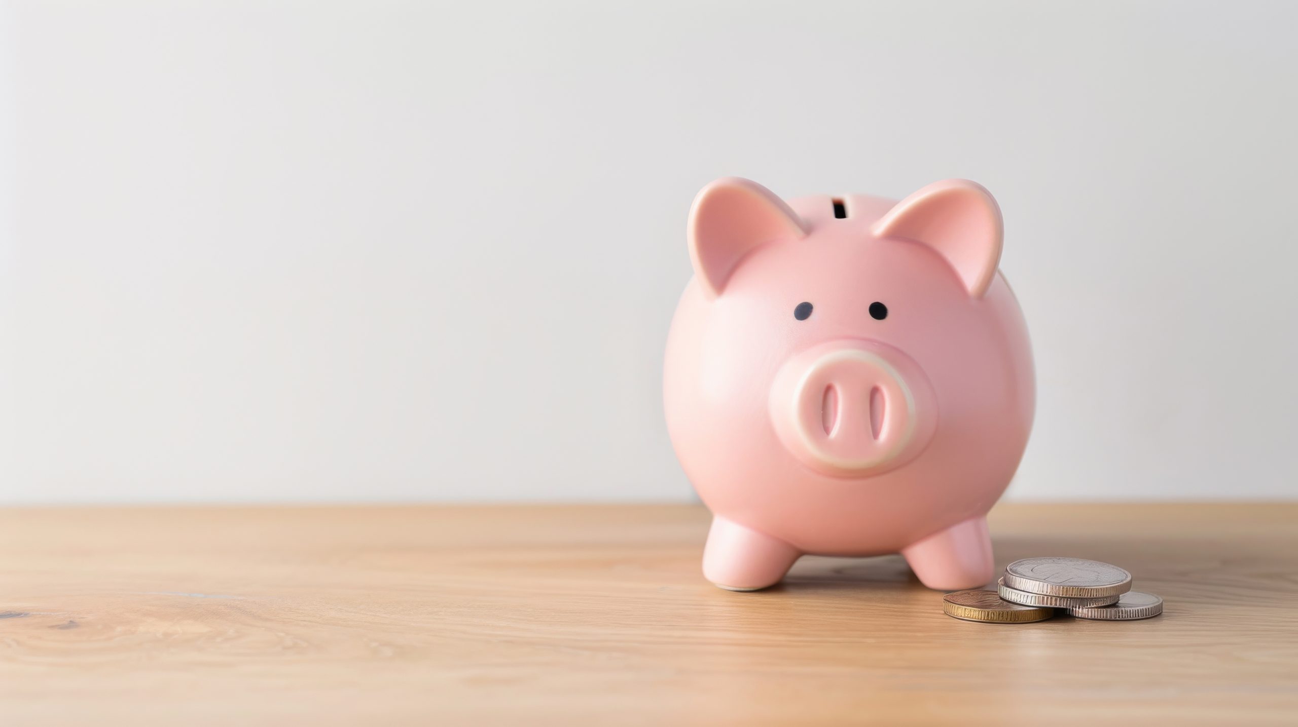 A pink piggy bank sits on a wooden surface with a small stack of coins beside it, against a plain white background.